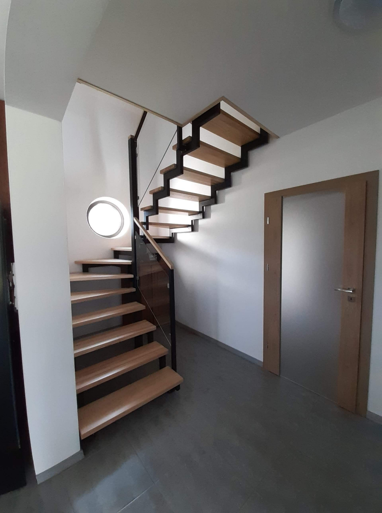 Nowoczesne schody drewniane z czarnymi metalowymi elementami i szklaną balustradą, oświetlone okrągłym oknem w ścianie, prowadzące do drzwi z matowego szkła.