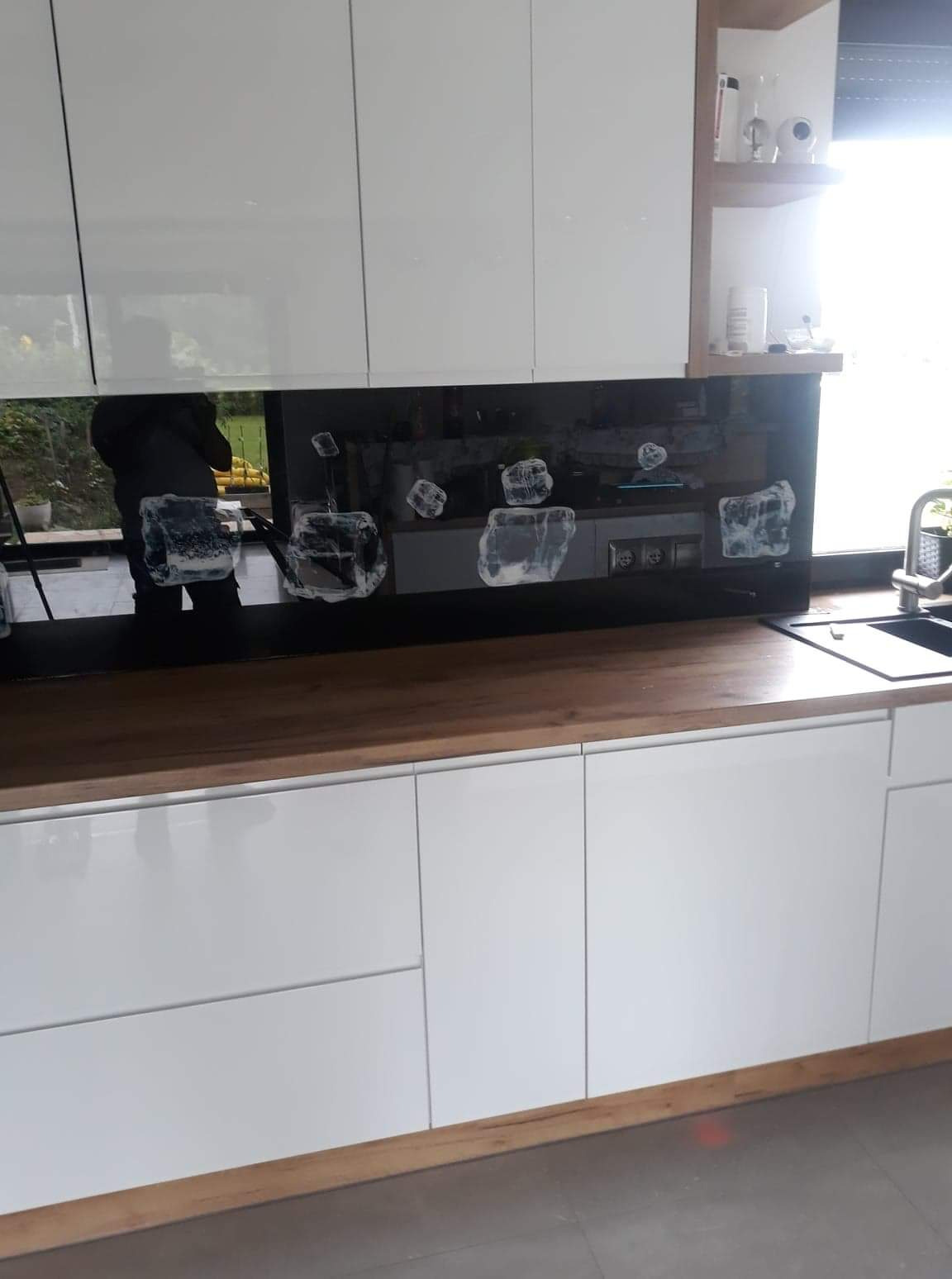 Szklany panel dekoracyjny z motywem geometrycznym, zamontowany nad blatem kuchennym z drewnianym wykończeniem, w nowoczesnej kuchni z białymi szafkami.