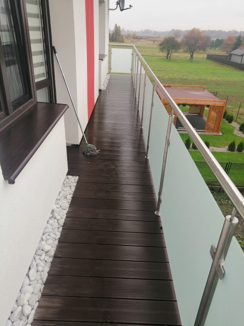 Balkon z ciemną drewnianą podłogą, białymi kamieniami przy ścianie i balustradą ze szkła matowego oraz stali nierdzewnej, z widokiem na ogród i altanę w tle, po umyciu podłogi mopem.