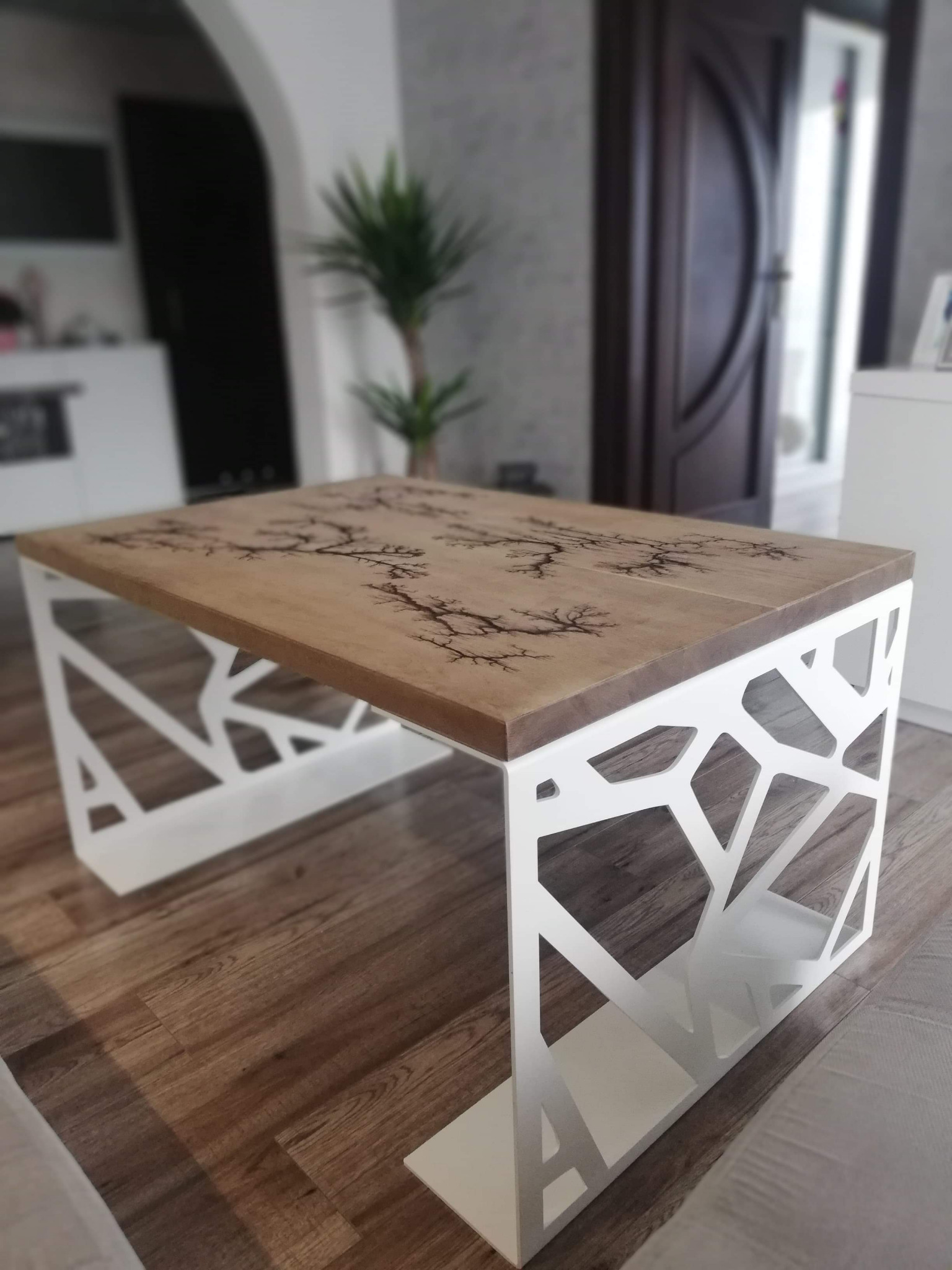 Stolik kawowy z drewnianym blatem zdobionym techniką Lichtenberga i ażurową, geometryczną podstawą z białego metalu.