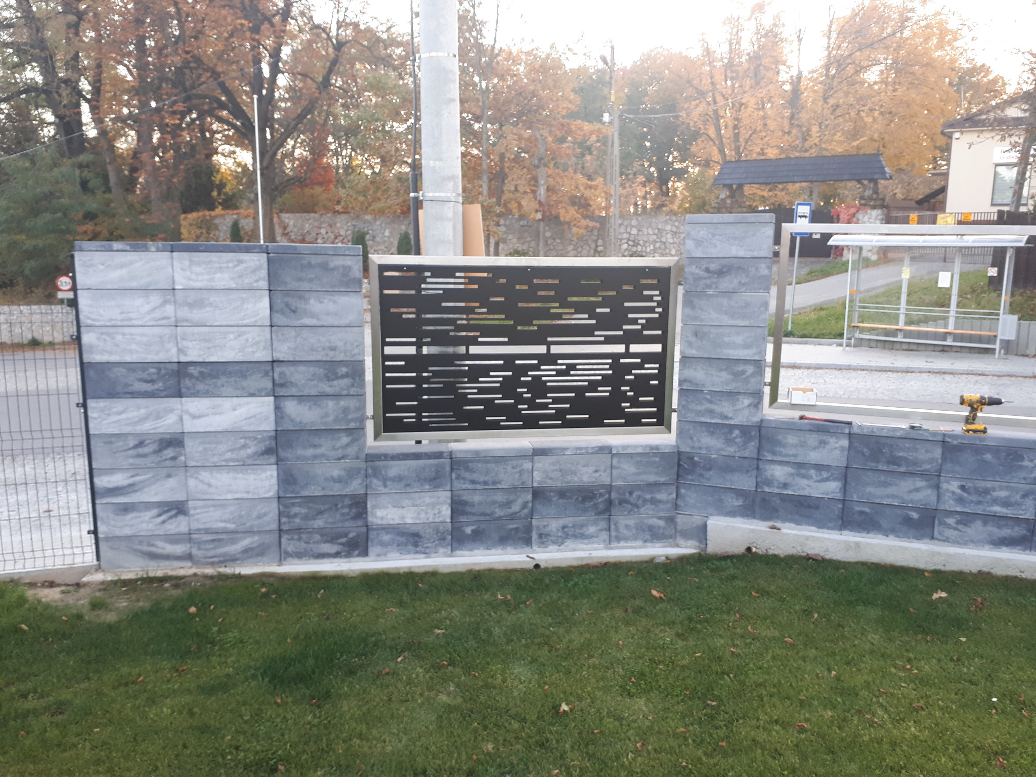 Nowoczesne ogrodzenie z szarej kostki betonowej z metalowym panelem o poziomym ażurowym wzorze, widoczne narzędzia na górze ogrodzenia, w tle przystanek autobusowy i drzewa.