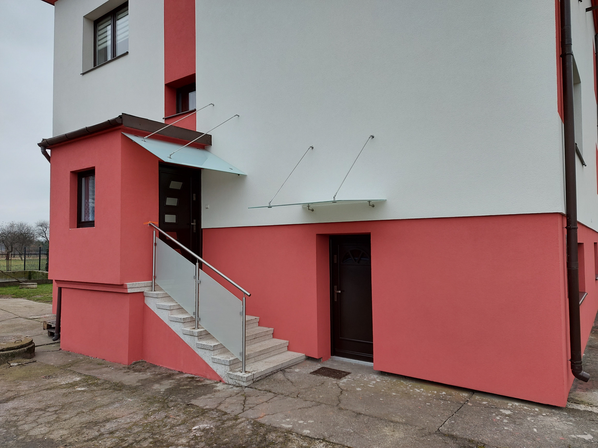 Zewnętrzne schody z jasnymi stopniami i balustradą ze szkła hartowanego prowadzące do drzwi wejściowych, w Skawinie, zadaszone przezroczystym daszkiem.