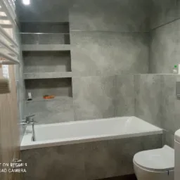 Nowoczesna łazienka z wanną obudowaną szarymi płytkami, wiszącą toaletą i grzejnikiem drabinkowym, z wnękami w ścianie pełniącymi rolę półek.