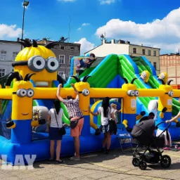 Duży, kolorowy dmuchany plac zabaw z motywem Minionków na miejskim placu, otoczony bawiącymi się dziećmi i ich rodzicami w słoneczny dzień.