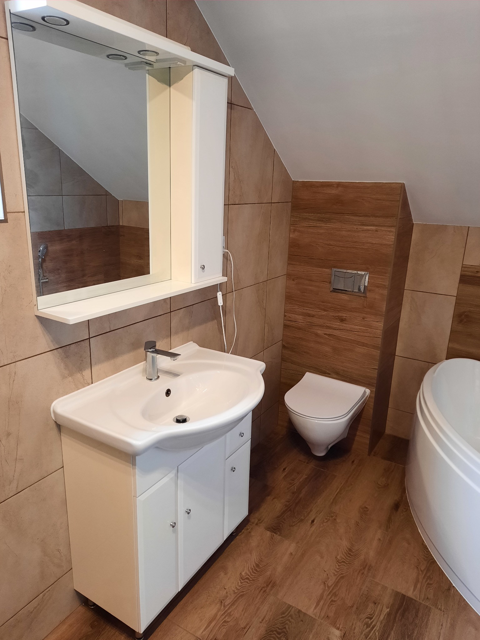 Ukończona łazienka na poddaszu z drewnopodobnymi płytkami na podłodze i ścianach, białą umywalką z szafką, lustrem z oświetleniem, podwieszanym WC i fragmentem wanny.