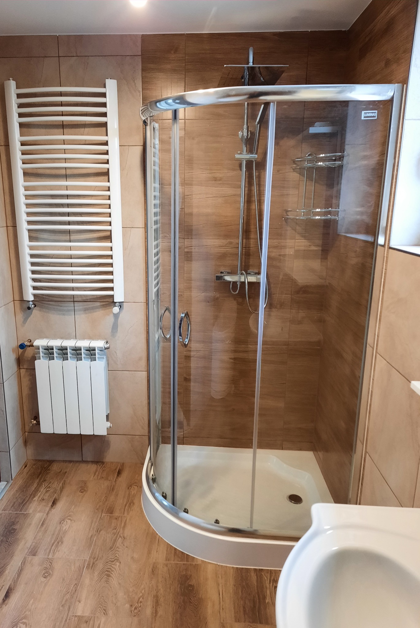 Wykończona łazienka z kabiną prysznicową narożną, grzejnikiem drabinkowym i klasycznym grzejnikiem, wykończona płytkami imitującymi drewno.