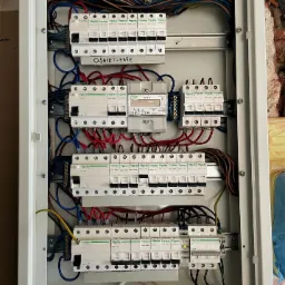 Otwarta skrzynka rozdzielcza Schneider Electric z bezpiecznikami, przewodami o różnych kolorach (niebieski, czerwony, brązowy, żółto-zielony) oraz licznikiem energii elektrycznej WZE-3, widoczne...