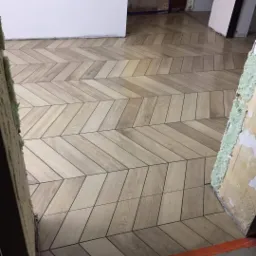 Ułożona w jodełkę jasna glazura imitująca drewno na podłodze, widoczne niedokończone wykończenia ścian.