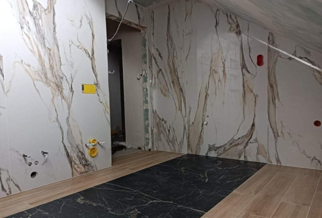 Wnętrze łazienki w trakcie prac wykończeniowych: ściany wyłożone płytkami imitującymi marmur z brązowymi żyłami, podłoga z płytek w dwóch odcieniach - czarnym i drewnopodobnym, widoczne...