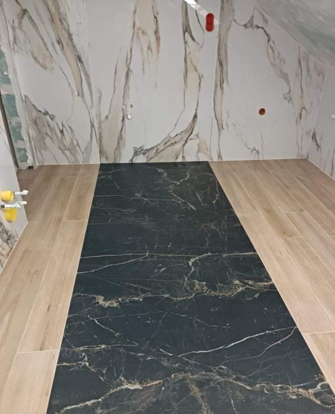 Wykończona łazienka na poddaszu z czarną marmurową wstawką pośrodku podłogi, obramowaną płytkami imitującymi jasne drewno, oraz ścianami wyłożonymi płytkami imitującymi biały marmur z brązowymi...