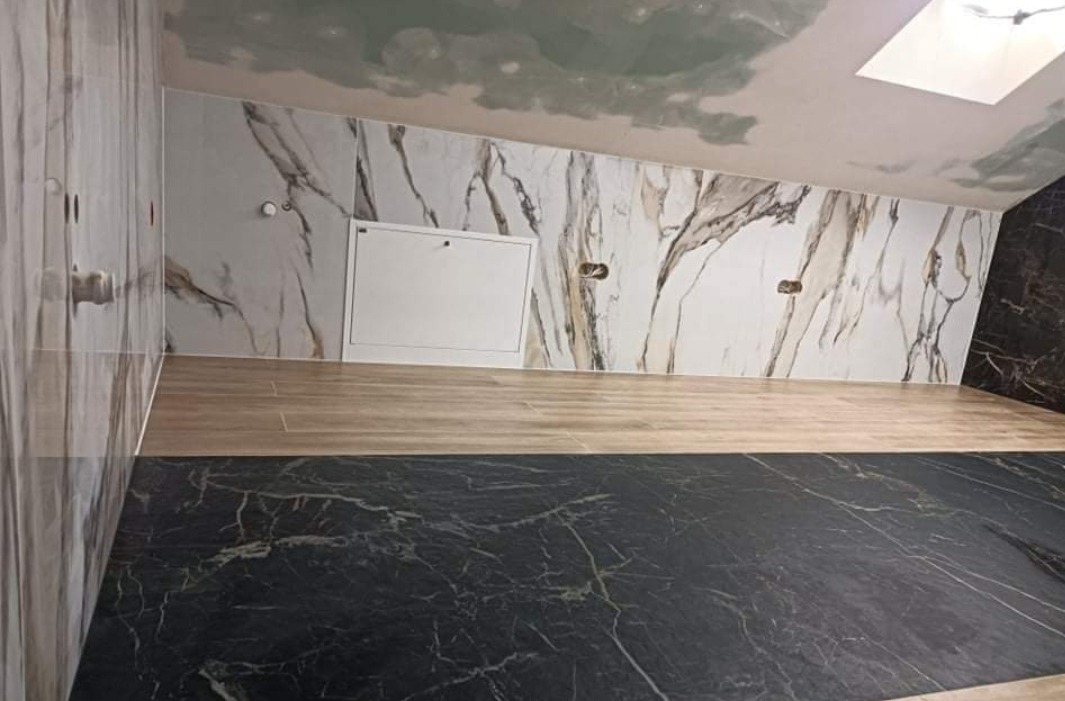 Wykończone wnętrze łazienki z marmurowymi płytkami na ścianach i podłodze, widoczny biały właz rewizyjny i skośny sufit z oknem dachowym.