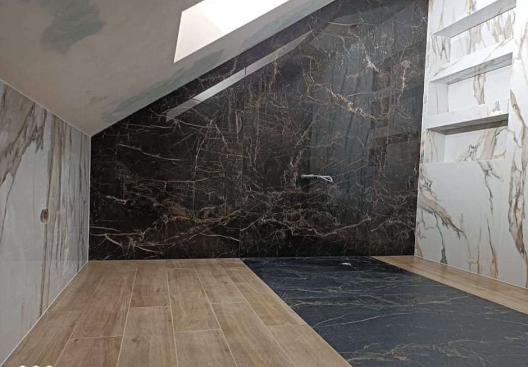 Ukończona łazienka na poddaszu z marmurowymi płytkami w odcieniach brązu i beżu na ścianach oraz podłodze, z drewnopodobnymi panelami podłogowymi w części pomieszczenia i wnękami w ścianie.