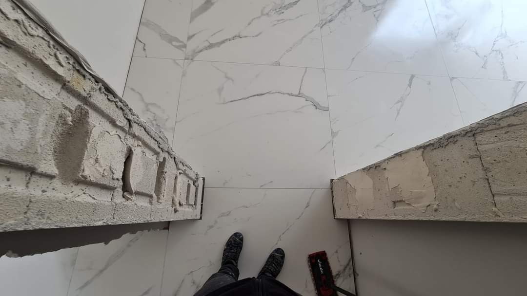 Białe płytki imitujące marmur na podłodze, widok z góry na fragment ściany z niedokończonymi zdobieniami z gipsu, czarne buty osoby fotografującej i czerwony mop.