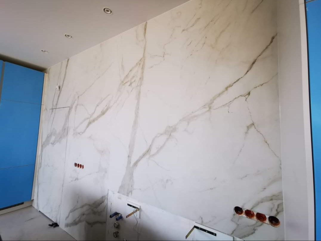 Ściana wyłożona wielkoformatowymi płytami imitującymi marmur Calacatta, widoczne otwory na gniazdka elektryczne i przyłącza wodno-kanalizacyjne, niebieska zabudowa meblowa po lewej stronie.