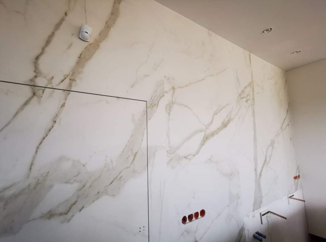 Ściana wykończona dużymi płytami imitującymi biały marmur z brązowymi żyłami, widoczne otwory na gniazdka elektryczne i rury wodne oraz czujnik ruchu.