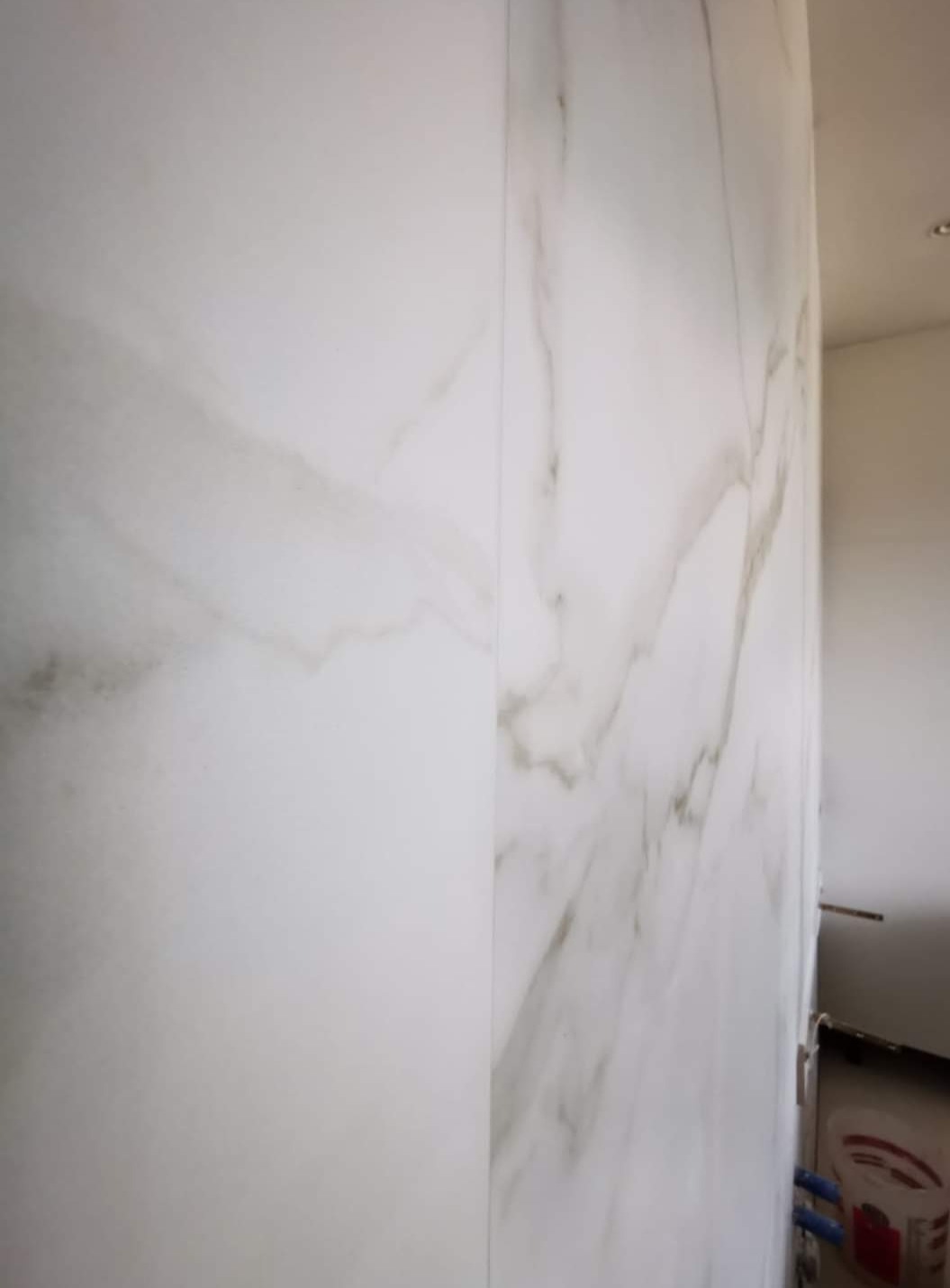 Ściana wyłożona dużymi, białymi płytami imitującymi marmur z delikatnymi szarymi żyłkami, widok z boku, fragment podłogi i instalacji hydraulicznej w tle.