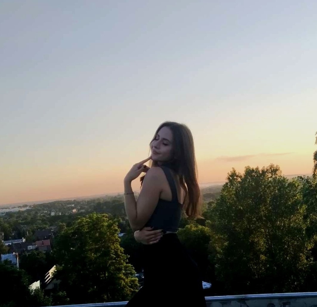 Portret młodej kobiety z długimi włosami, w ciemnym topie i spódnicy, stojącej na tle panoramy miasta o zmierzchu, z delikatnym uśmiechem i ręką przy twarzy.