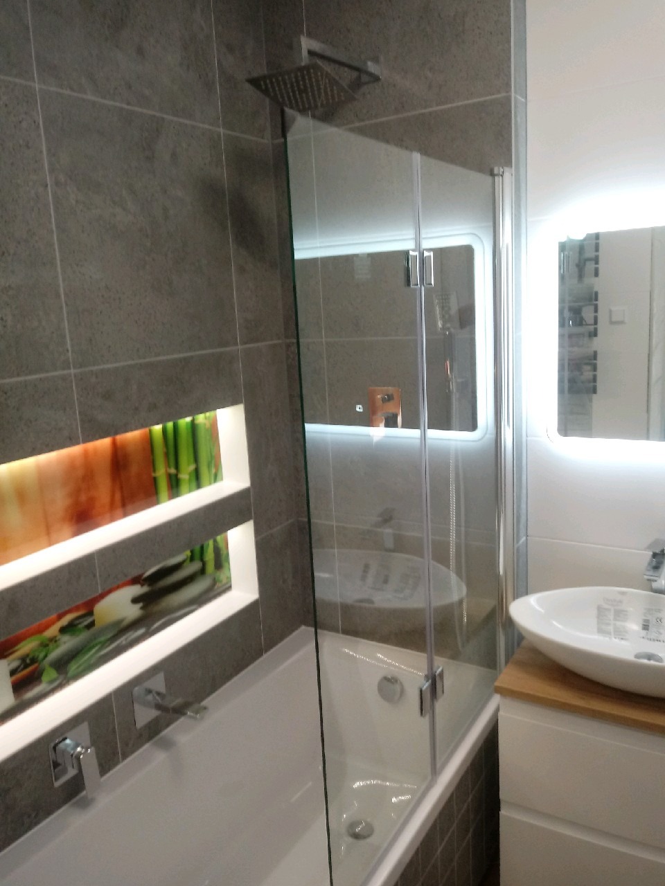 Nowoczesna łazienka z szarymi płytkami, kabiną nawannową z przezroczystego szkła, podświetlanym lustrem i dekoracyjną wnęką z motywem roślinnym.