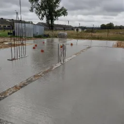 Świeżo wylana, mokra płyta fundamentowa z wystającymi prętami zbrojeniowymi, pomarańczowe rury wentylacyjne widoczne w betonie, pochmurne niebo w tle.