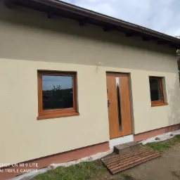 Świeżo otynkowany, beżowy budynek z brązowymi oknami i drzwiami, widoczne fragmenty instalacji elektrycznej wystające ze ściany, zabezpieczenia folią malarską u podstawy elewacji.