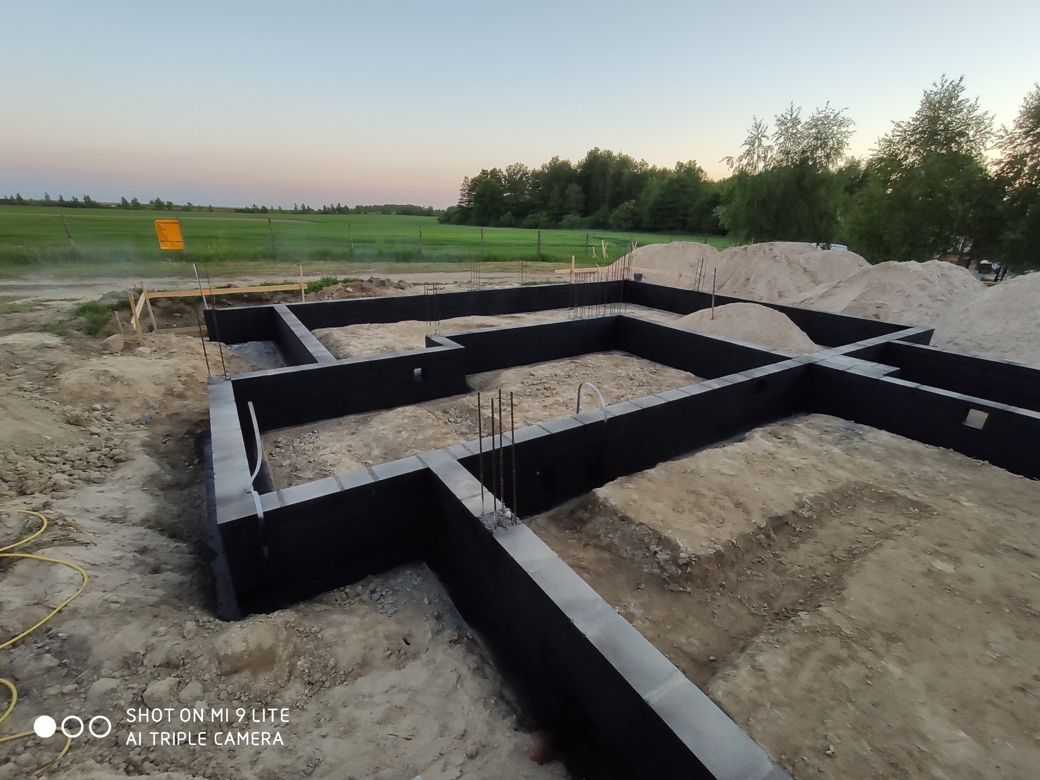 Fundamenty domu jednorodzinnego wykonane z bloczków betonowych, zabezpieczone izolacją pionową, widoczne pręty zbrojeniowe, w tle hałdy piasku i zielone pola uprawne pod wieczornym niebem.