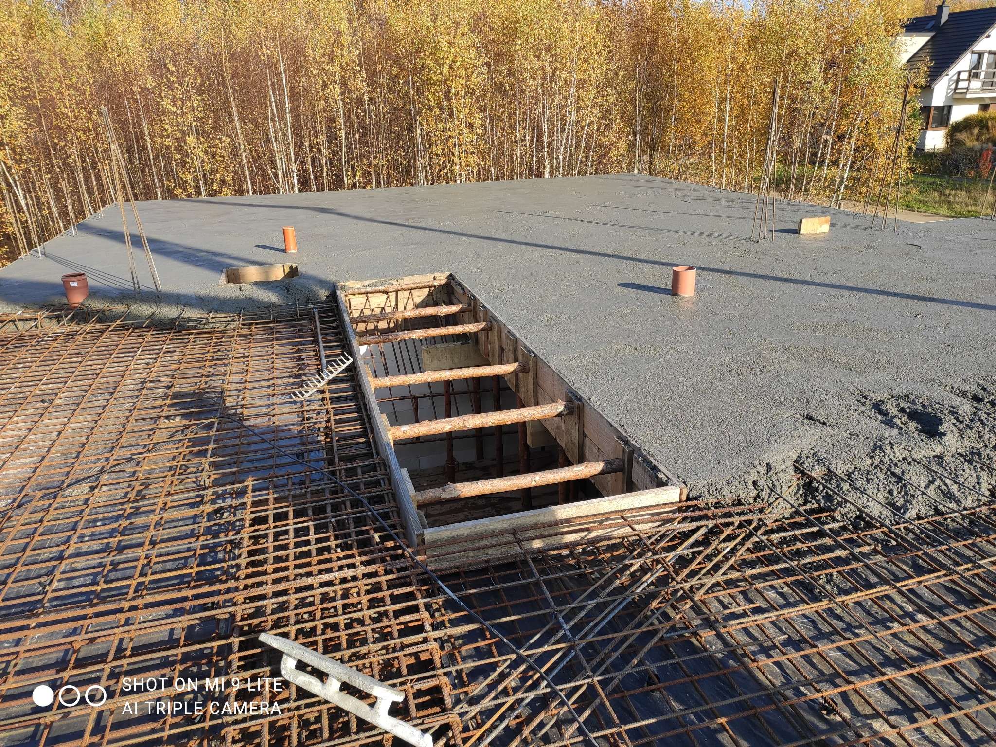Zalany betonem strop z widocznym zbrojeniem i otworem szalunkowym, w tle drzewa z żółtymi liśćmi i fragment budynku.