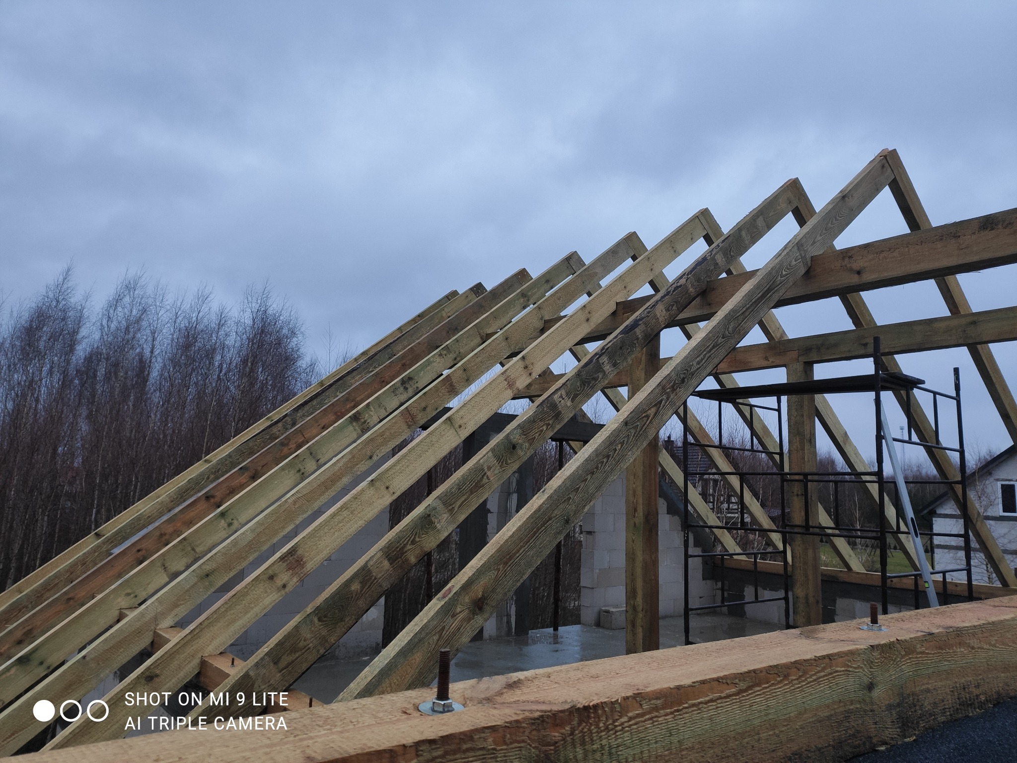 Konstrukcja drewniana dachu w trakcie budowy, widoczne krokwie i murłaty, w tle budynek z białego pustaka i pochmurne niebo.