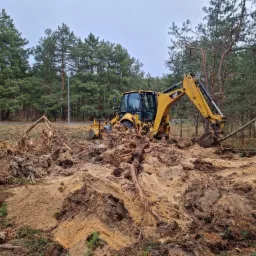 Żółta koparko-ładowarka CAT usuwa korzenie i pnie drzew na zalesionym terenie, przygotowując grunt pod budowę.
