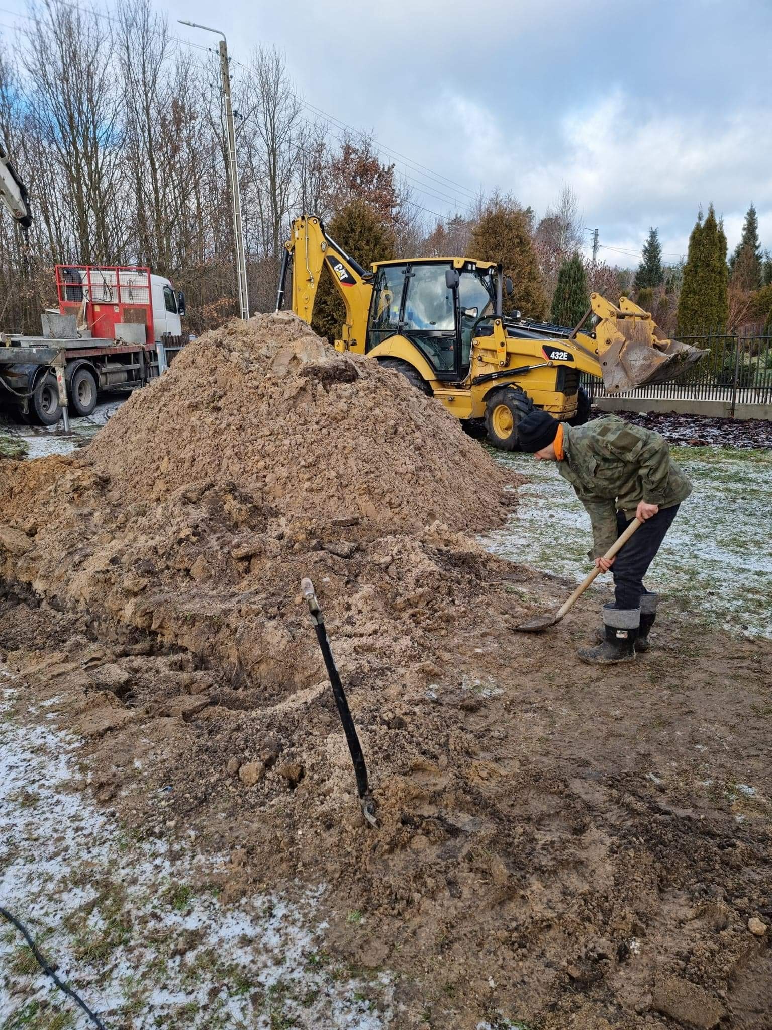 Mężczyzna w wojskowej kurtce kopie łopatą ziemię obok koparki CAT 432E i ciężarówki, na częściowo ośnieżonym terenie.