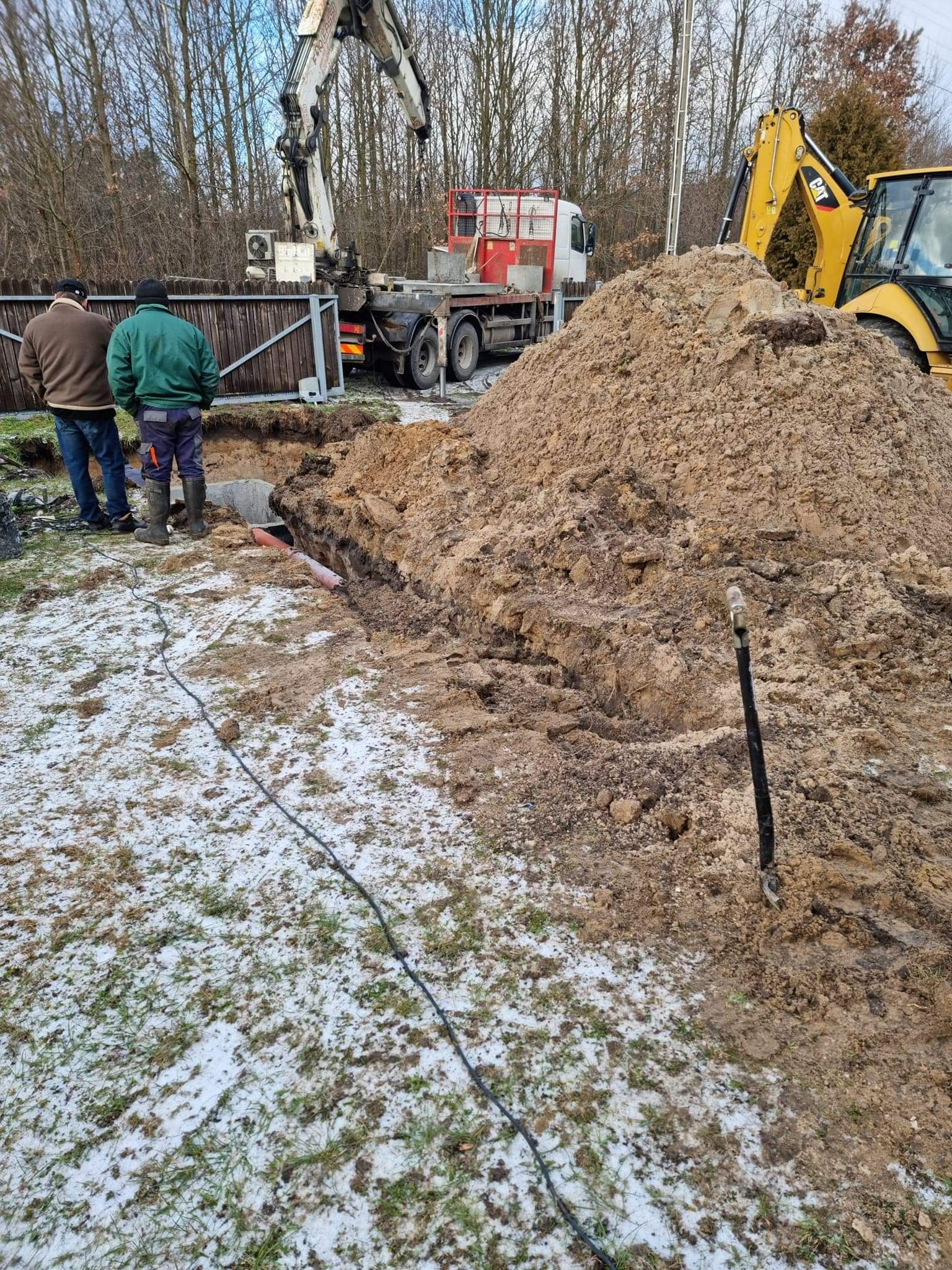 Wykop pod fundament z widocznymi rurami, stertą ziemi, koparką CAT i dźwigiem na tle ośnieżonej trawy i drzew.