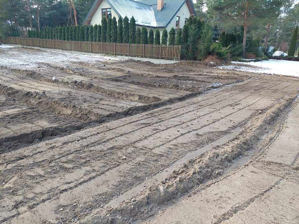 Przygotowany teren pod budowę domu, widoczne ślady po maszynach, w tle dom jednorodzinny i rząd tuj.