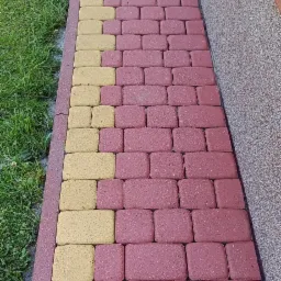 Ułożona kostka brukowa w kolorze czerwonym i żółtym, tworząca ścieżkę w Lublinie, przylegająca do trawnika i ściany budynku z widocznym parapetem.