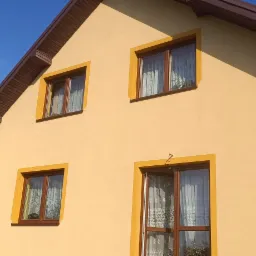 Żółta elewacja domu z brązowym dachem i oknami z żółtymi ramami, widok z dołu na bocznej ścianie. Na dachu panele słoneczne.