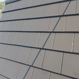 Simon Wash - Czysty, szary dach z ceramicznej dachówki w słoneczny dzień. Widoczny cień osoby w lewym dolnym rogu. Dach po renowacji lub świeżo położony.