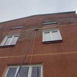 Simon Wash - Elewacja budynku z widoczną różnicą czystości po umyciu fragmentu, z użyciem teleskopowej lancy. Widoczne okna i struktura tynku.