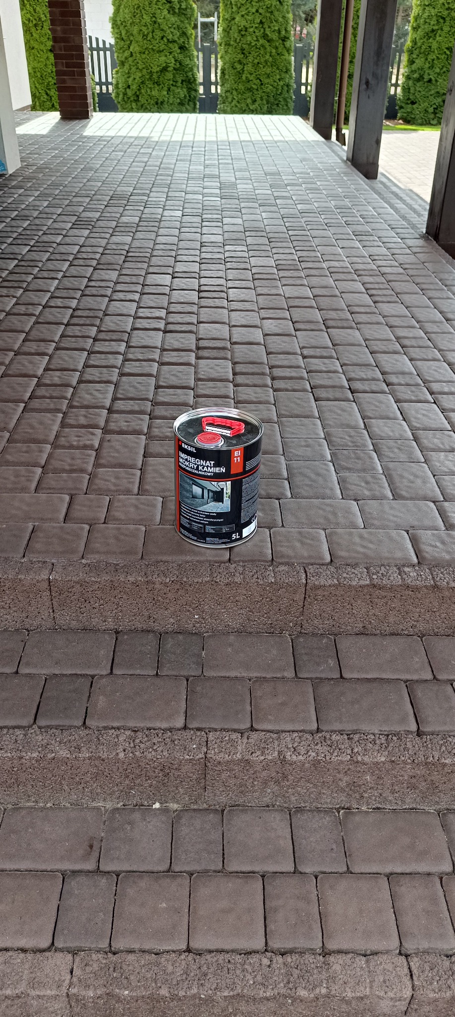 Puszka impregnatu do kostki brukowej Eksil EI 11 na tle wyczyszczonej, brązowej kostki brukowej przed budynkiem z kolumnami i tujami w tle.