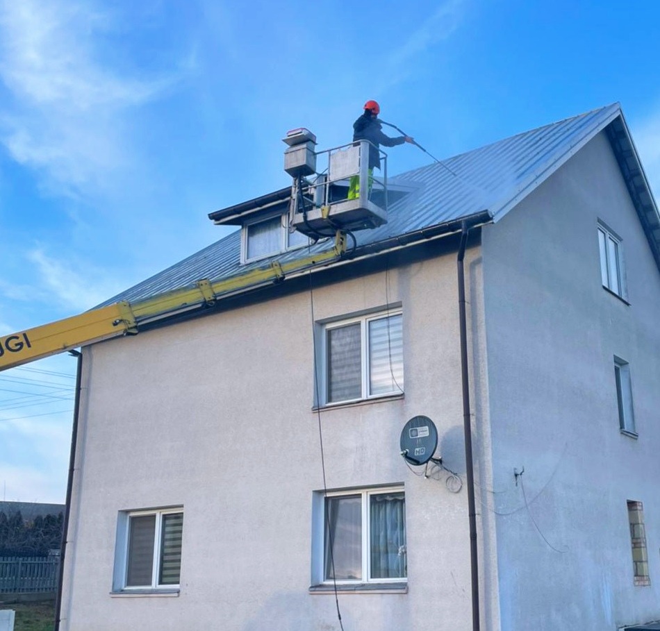 Pracownik w pomarańczowym kasku na podnośniku koszowym czyści dach dwupiętrowego domu strumieniem wody pod ciśnieniem.