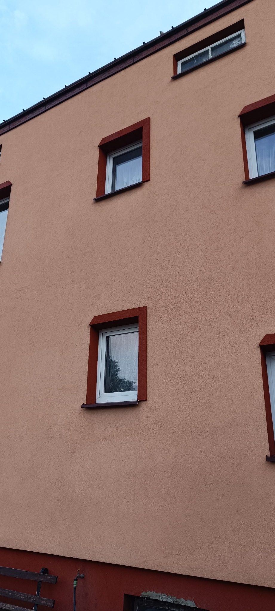Fragment elewacji budynku z oknami w brązowych ramach, widoczne z bliska, z teksturą tynku baranka w odcieniach beżu.