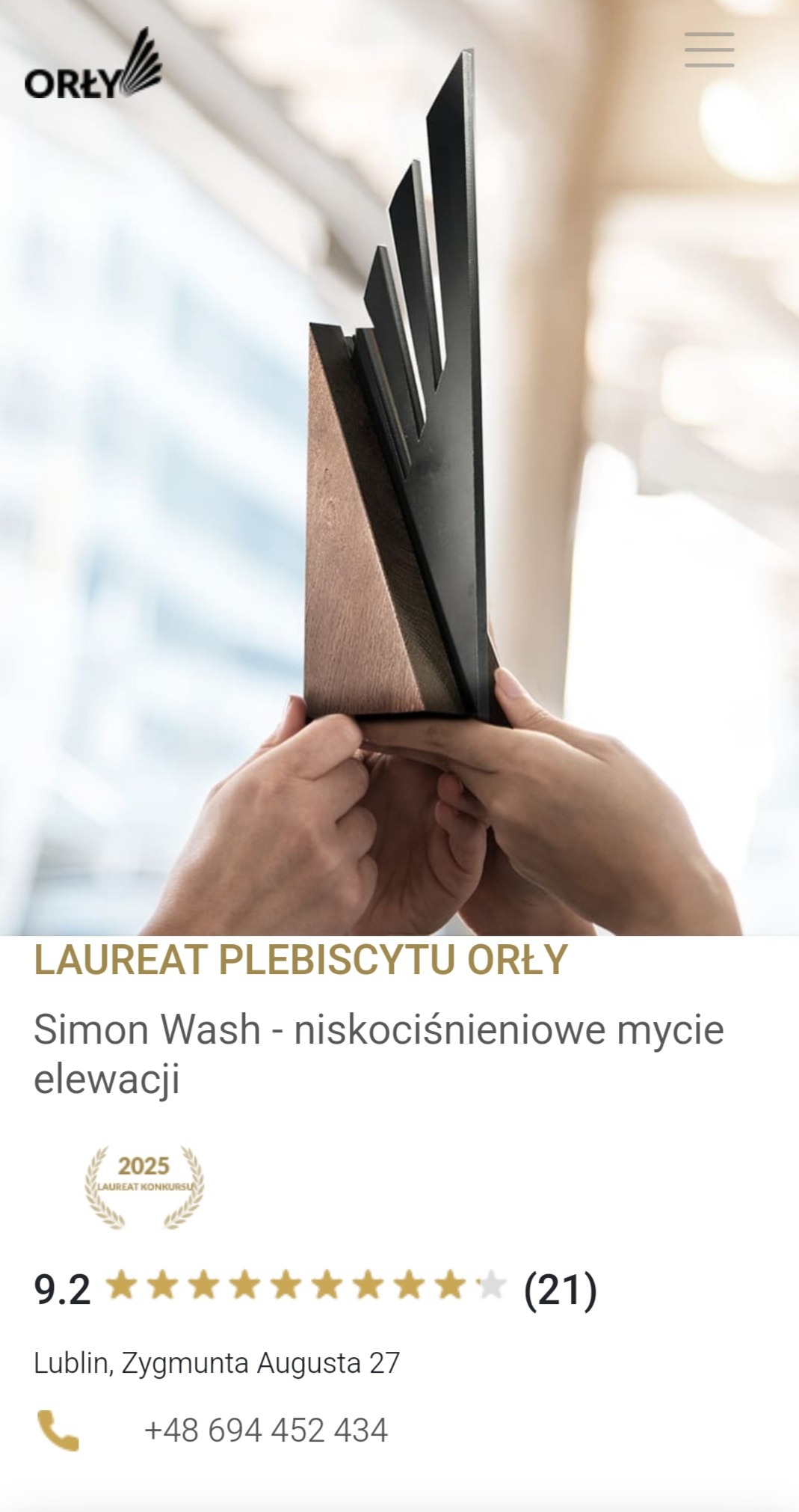 Statuetka nagrody 'Orły' za niskociśnieniowe mycie elewacji, trzymana w dłoniach na rozmytym tle biurowym.