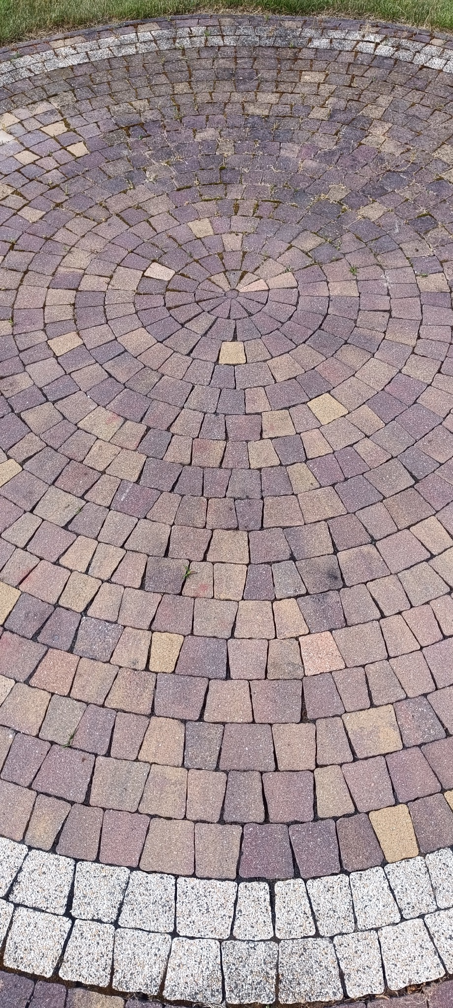 Ułożona kostka brukowa w okrągły wzór, z widocznymi różnymi odcieniami brązu i beżu, otoczona obrzeżem z jasnej kostki. Widoczne źdźbła trawy między kostkami.