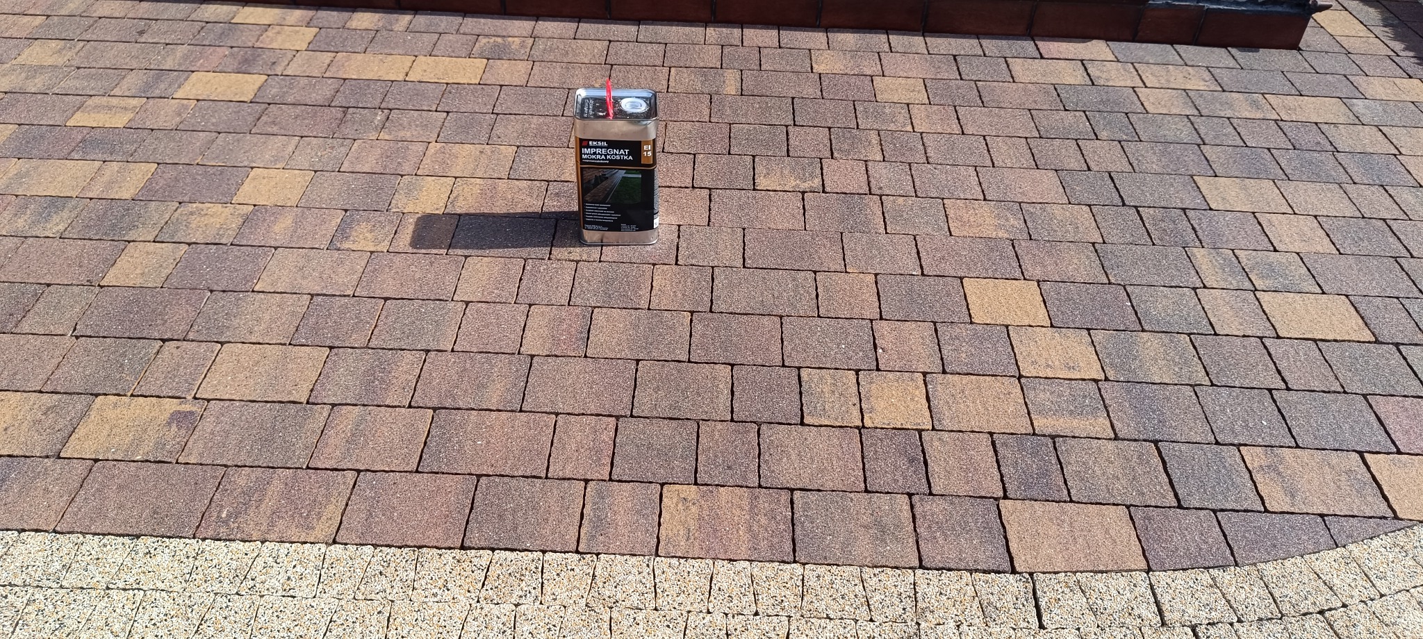 Impregnat do kostki brukowej na tle ułożonej kostki w odcieniach brązu i beżu. Widoczna faktura kostki i granica z jaśniejszym, drobnym brukiem.