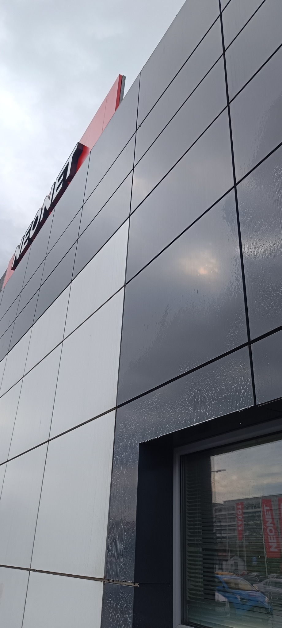 Fragment elewacji budynku z napisem NEONET, część paneli umyta, widać spływającą wodę, okno z odbiciem budynku. Ujęcie z dołu.