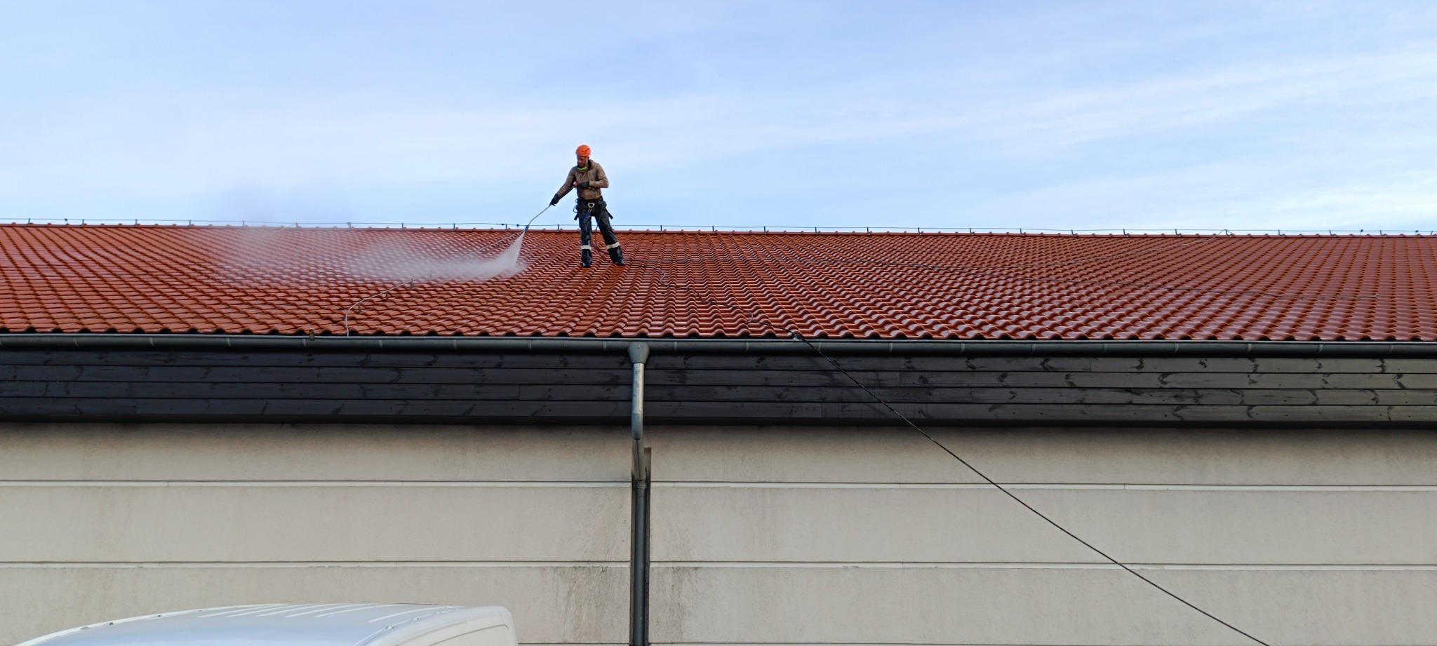 Pracownik w kasku myje czerwoną dachówkę myjką ciśnieniową. Widoczny strumień wody i czyszczona powierzchnia dachu. Praca na wysokościach, zabezpieczenia linowe.