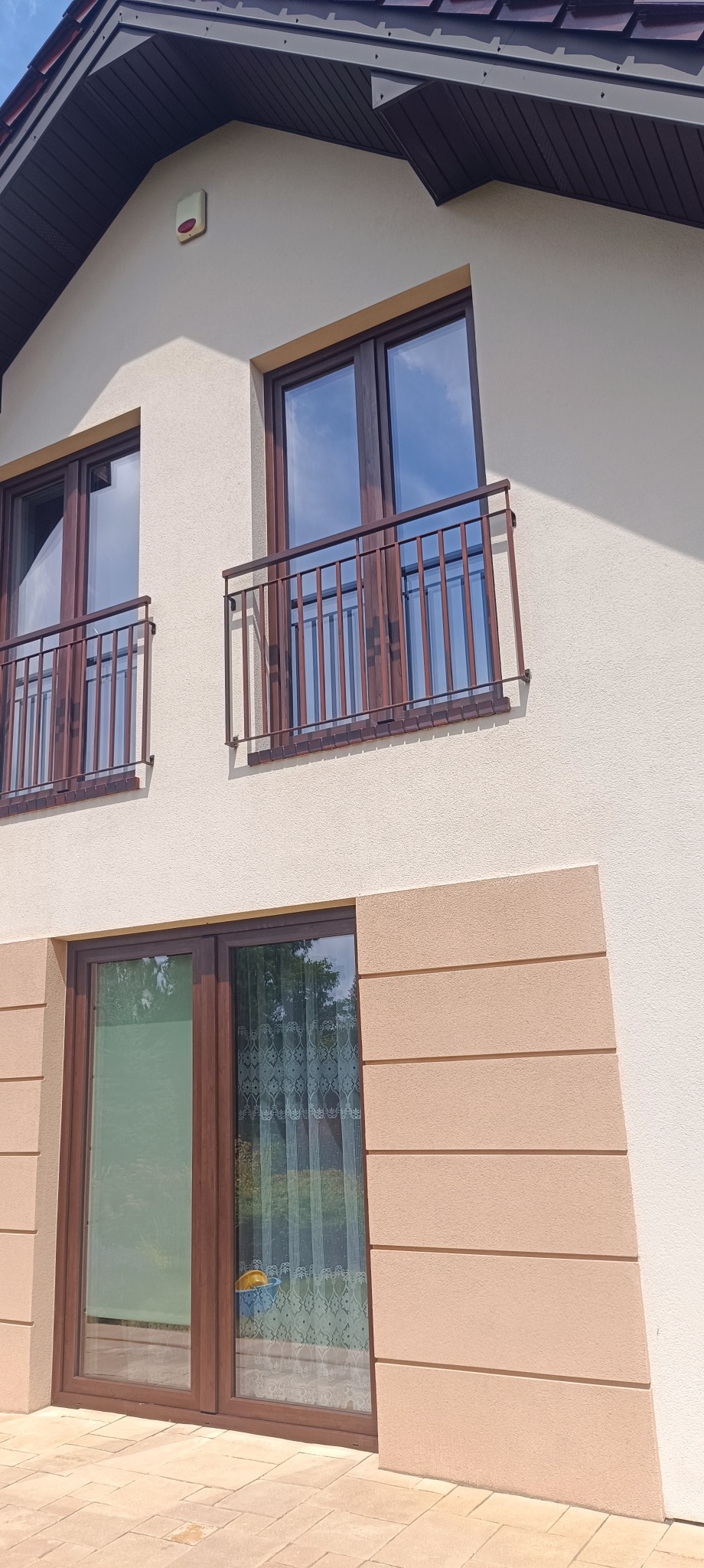 Jasna elewacja domu z brązowymi oknami i balkonami. Detal: dekoracyjne pasy w dolnej części fasady. Widoczne odbicie nieba w szybach okien.