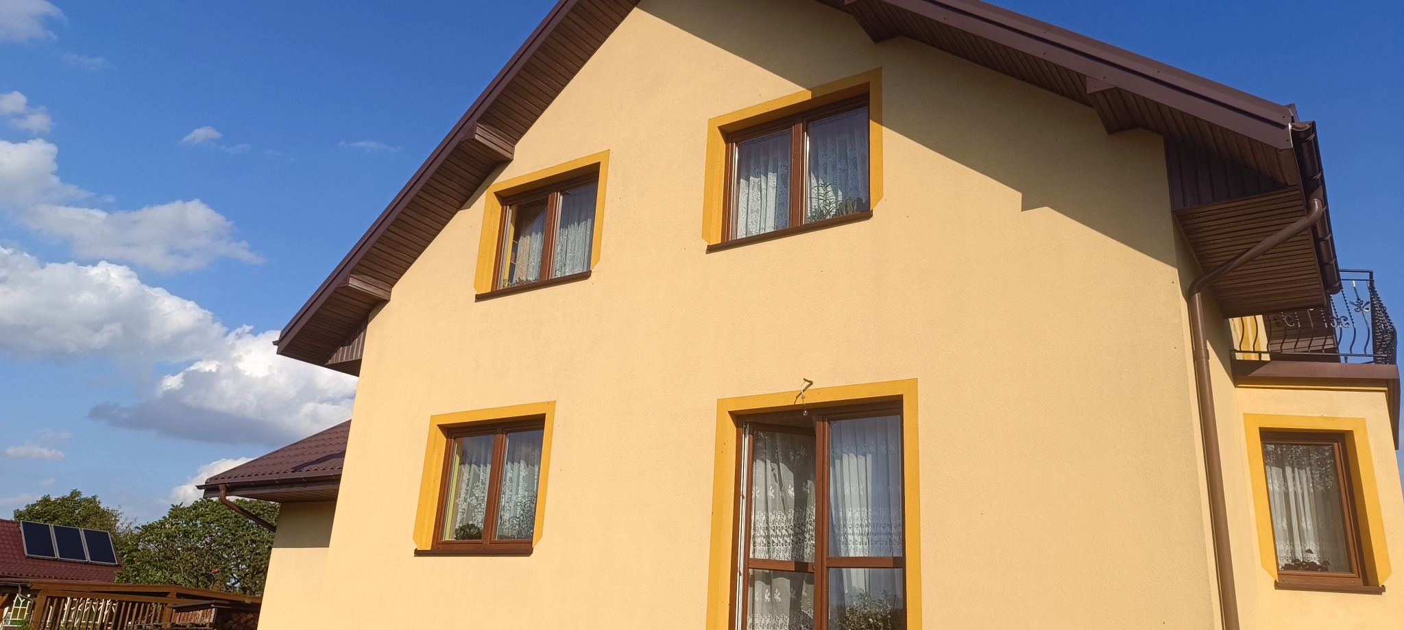 Żółta elewacja domu z brązowym dachem i oknami z żółtymi ramami, widok z dołu na bocznej ścianie. Na dachu panele słoneczne.