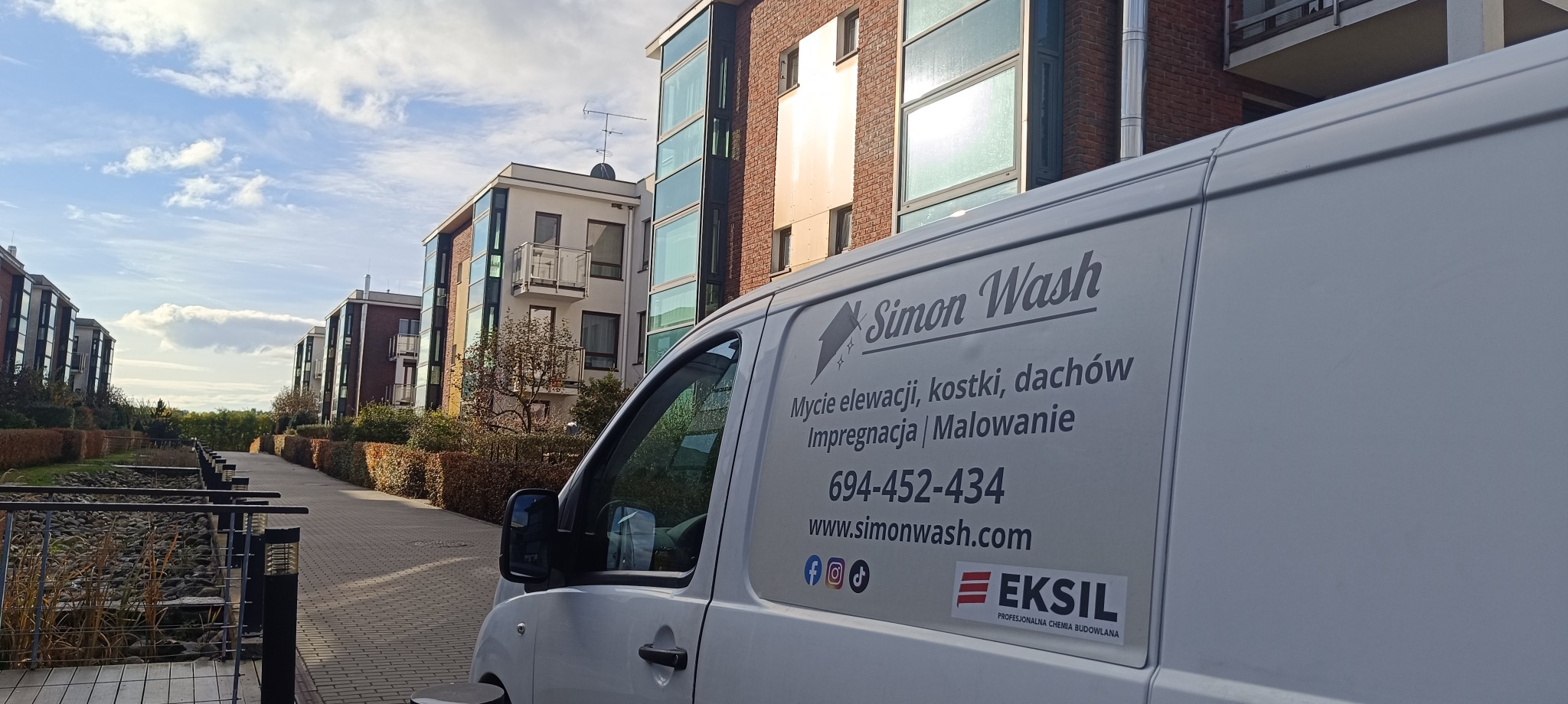 Firmowy bus z logo Simon Wash na tle nowoczesnych budynków. Oferta: mycie elewacji, kostki, dachów, impregnacja i malowanie. Kontakt: 694-452-434, simonwash.com.