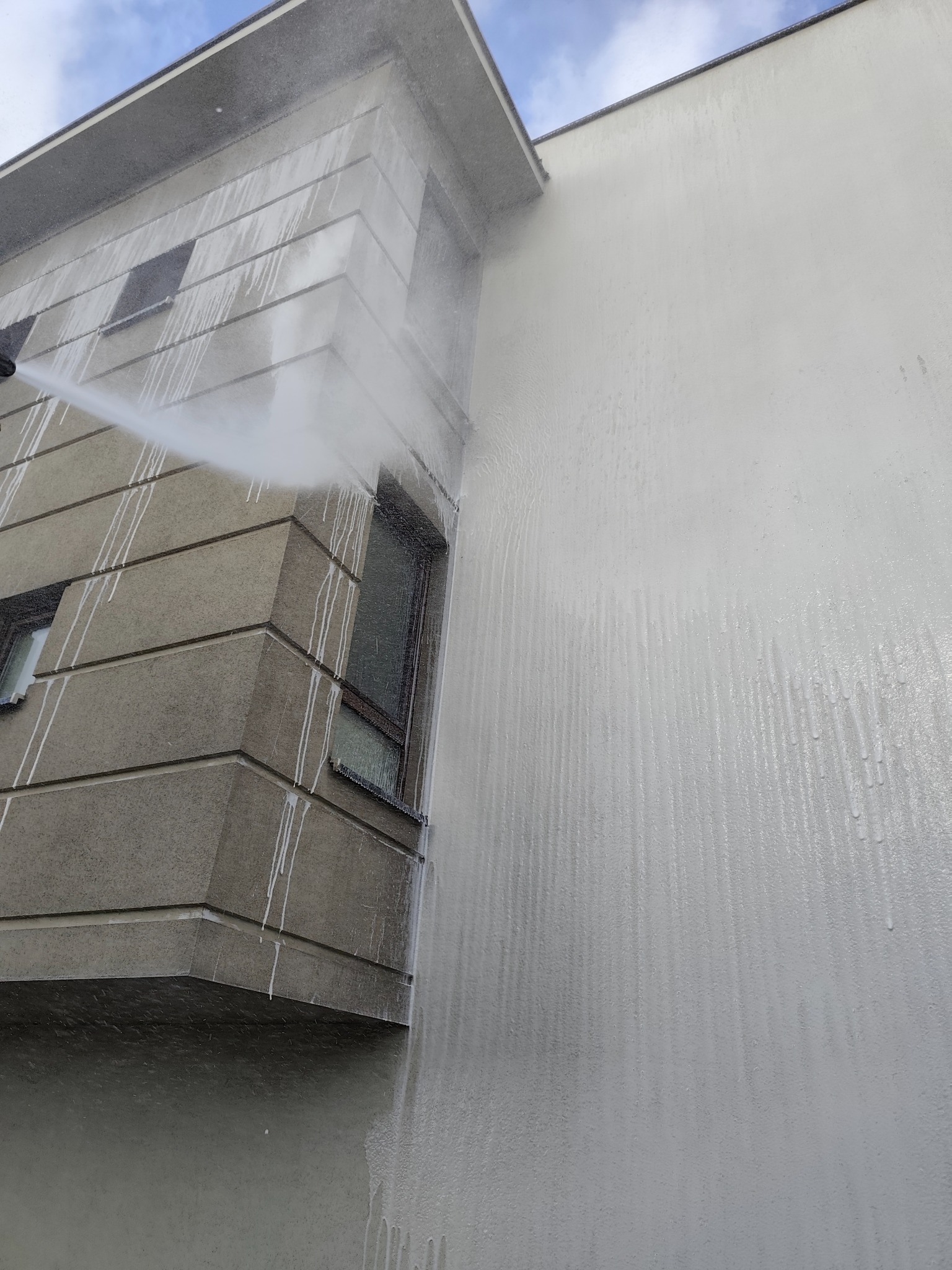 Intensywne strumienie wody pod ciśnieniem usuwają zabrudzenia z elewacji budynku, ukazując efekt czyszczenia na jasnej, zewnętrznej ścianie.