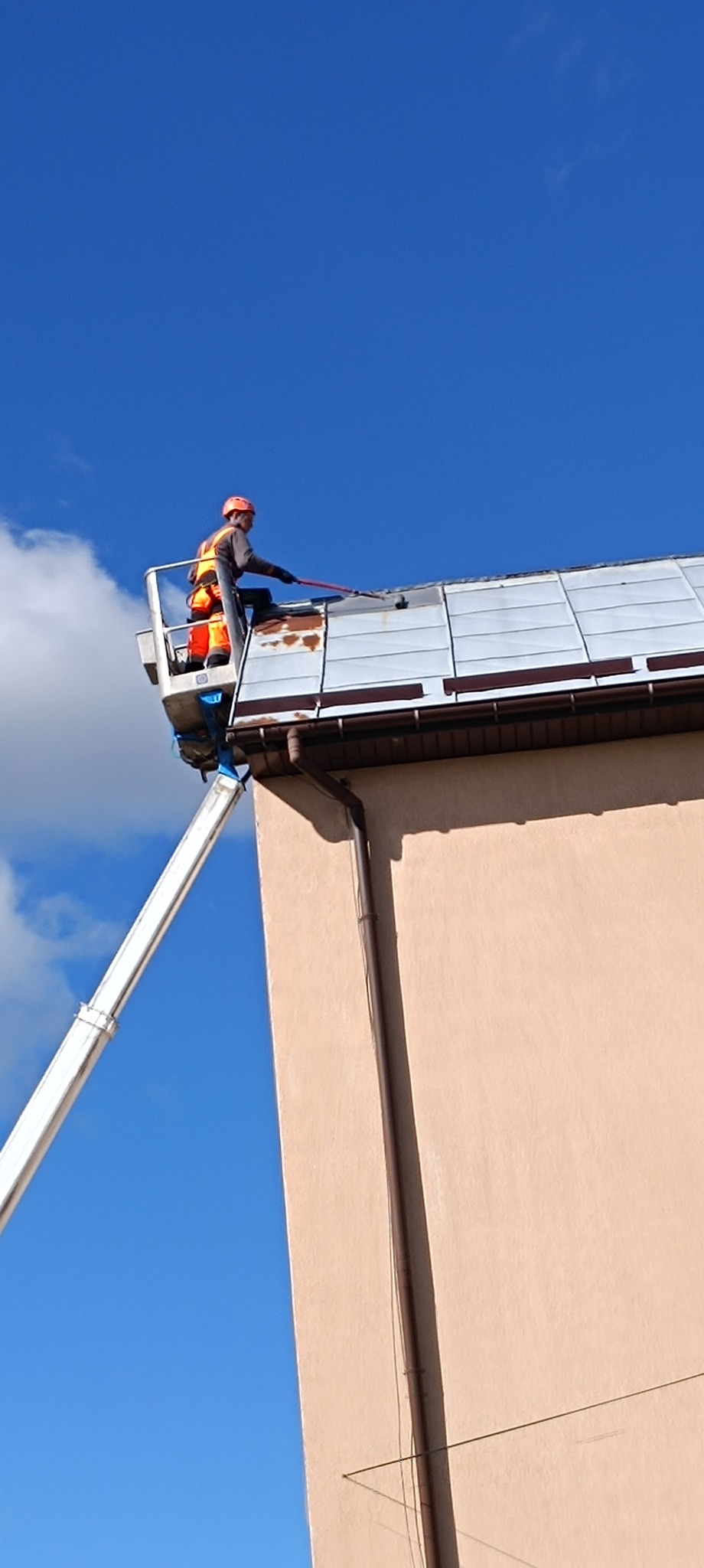 Pracownik w pomarańczowym kombinezonie na podnośniku czyści dach budynku za pomocą długiej szczotki. Błękitne niebo w tle.
