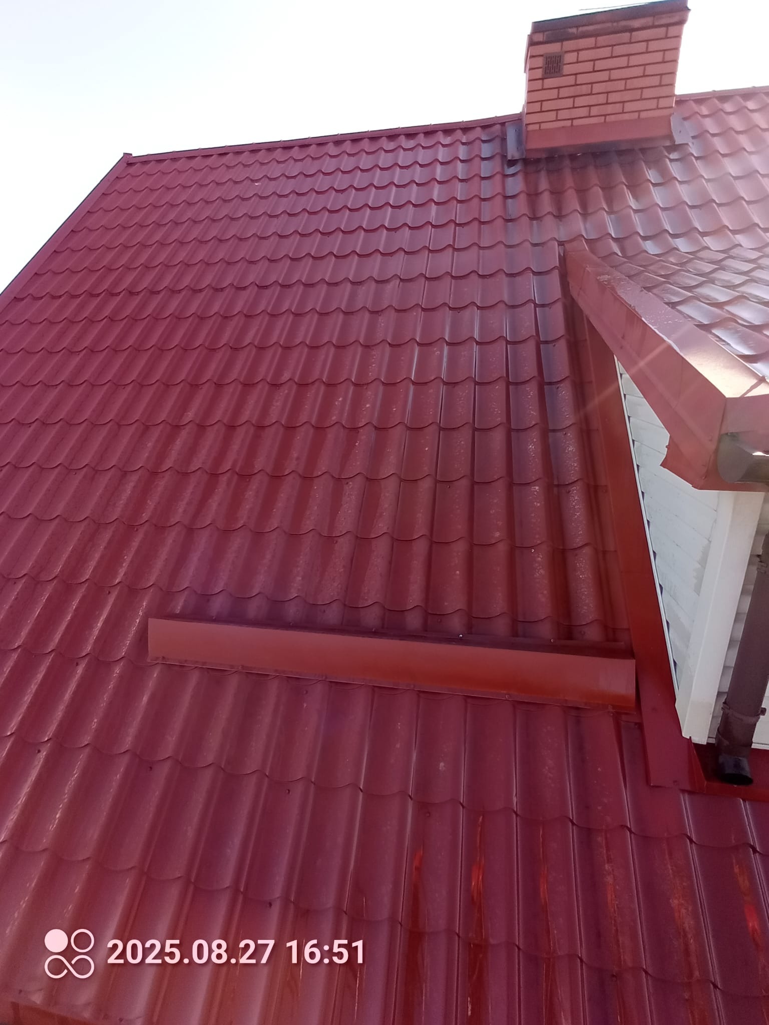 Czerwony dach z blachy trapezowej z ceglanym kominem w słoneczny dzień. Widoczne ślady zabrudzeń na powierzchni dachu. Ujęcie z dołu, lekko z boku.