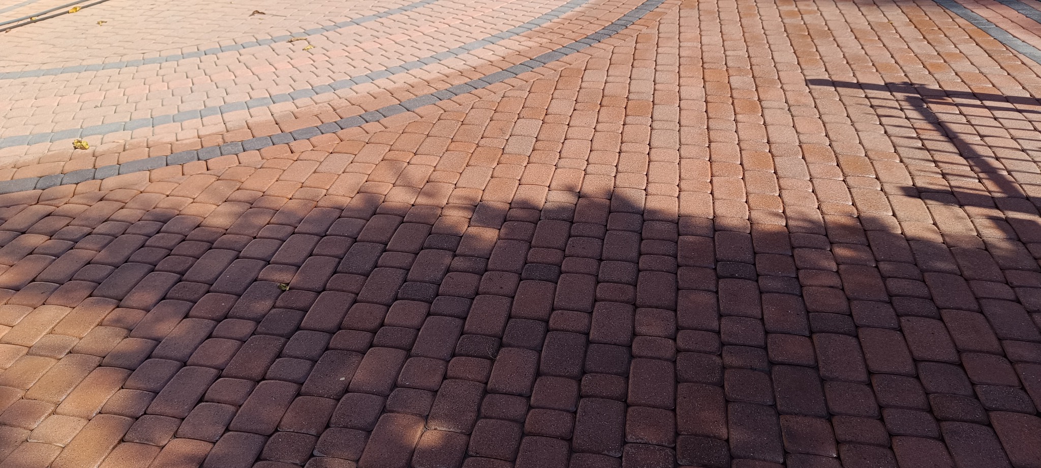 Ułożona, brązowa kostka brukowa z szarymi obrzeżami, częściowo zacieniona, z widocznym cieniem słupka. Układ w jodełkę, regularny wzór.